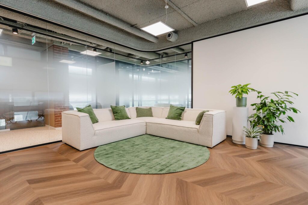 Modern office lounge area with a white corner sofa, green cushions, a round green rug, and potted plants on a wooden herringbone floor.